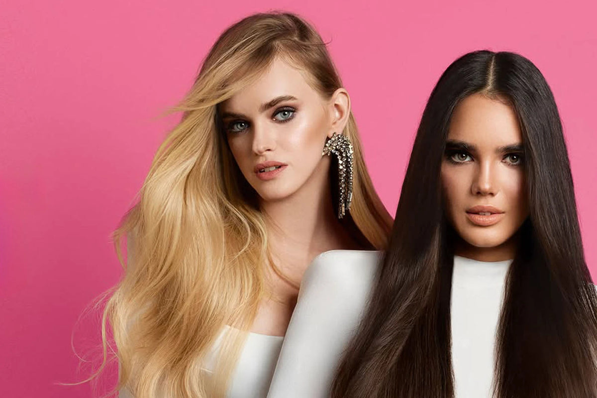 Two models displaying long, flowing Seamless1 professional hair extensions demonstrating seamless blend and premium quality on pink backdrop